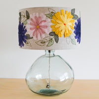 A hand-embroidered lampshade with a floral pattern in navy blue, sunny yellow, and rosy pink, displayed on a glass lamp base.
