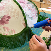 A close-up of a person's hands using a blue hot glue gun to apply adhesive to green fringe trim on a dome-shaped lampshade with a pink and green floral pattern.