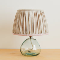 A natural linen gathered lampshade with a pink gathered texture, placed on a wooden surface with a glass lamp base.