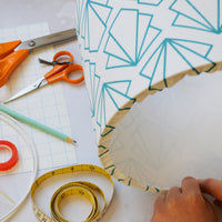 A person crafting a lampshade with geometric patterns, sitting at a table with scissors, tape measure, and other materials.