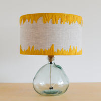 A yellow edged embroidered lampshade with a linen base, resting on a glass lamp base.