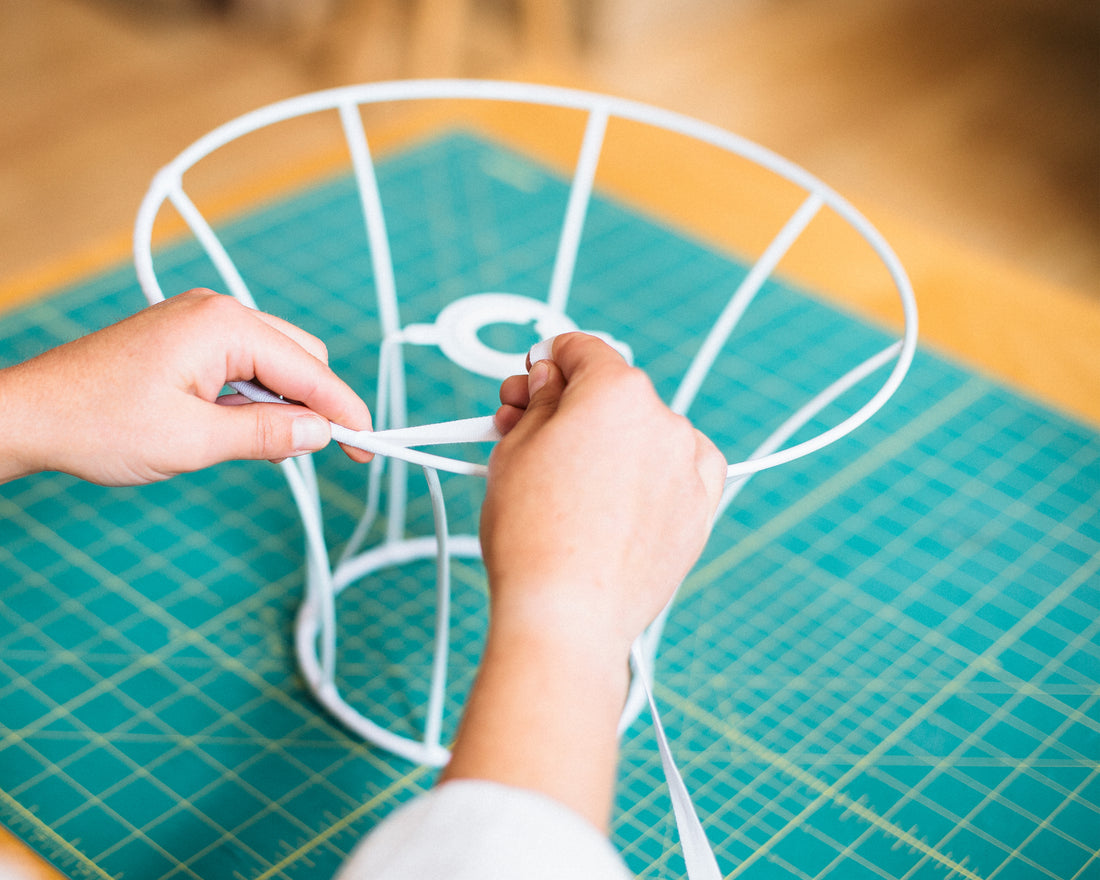 A person's hands binding a lampshade frame with white cotton tape.