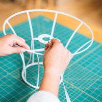A person's hands binding a lampshade frame with white cotton tape.