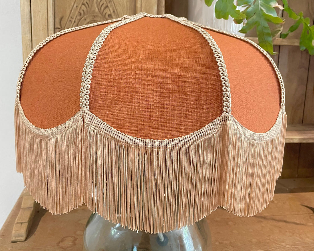 A dome-shaped table lamp shade in terracotta orange fabric with a beige fringe trim and bead detailing, displayed on a clear glass base on a wooden table.