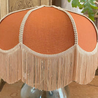 A dome-shaped table lamp shade in terracotta orange fabric with a beige fringe trim and bead detailing, displayed on a clear glass base on a wooden table.