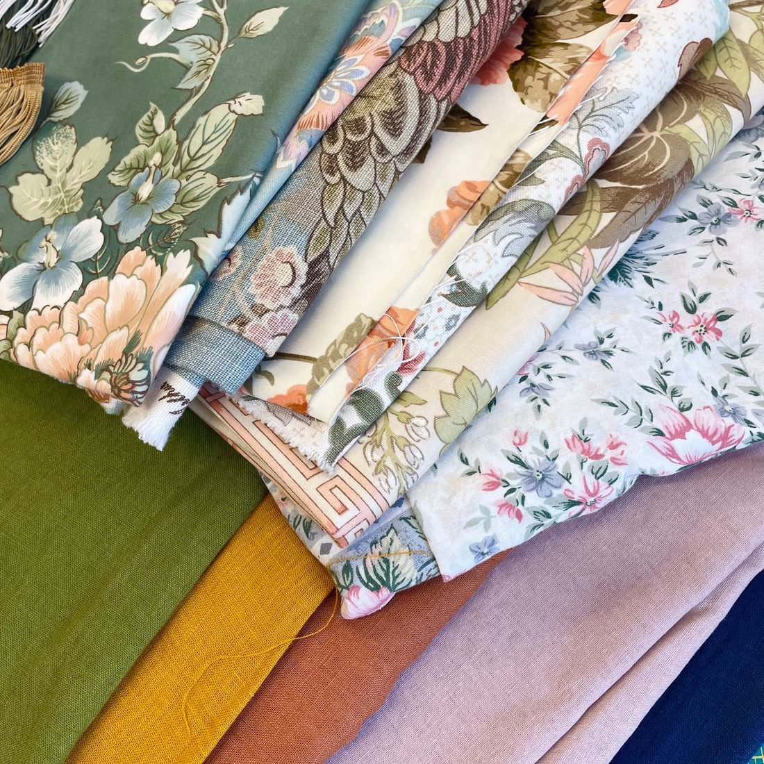 A stack of folded patterned fabrics in various colors and designs, including floral, bird, and abstract prints, arranged on a cutting mat, showing the fabric selection in the workshop