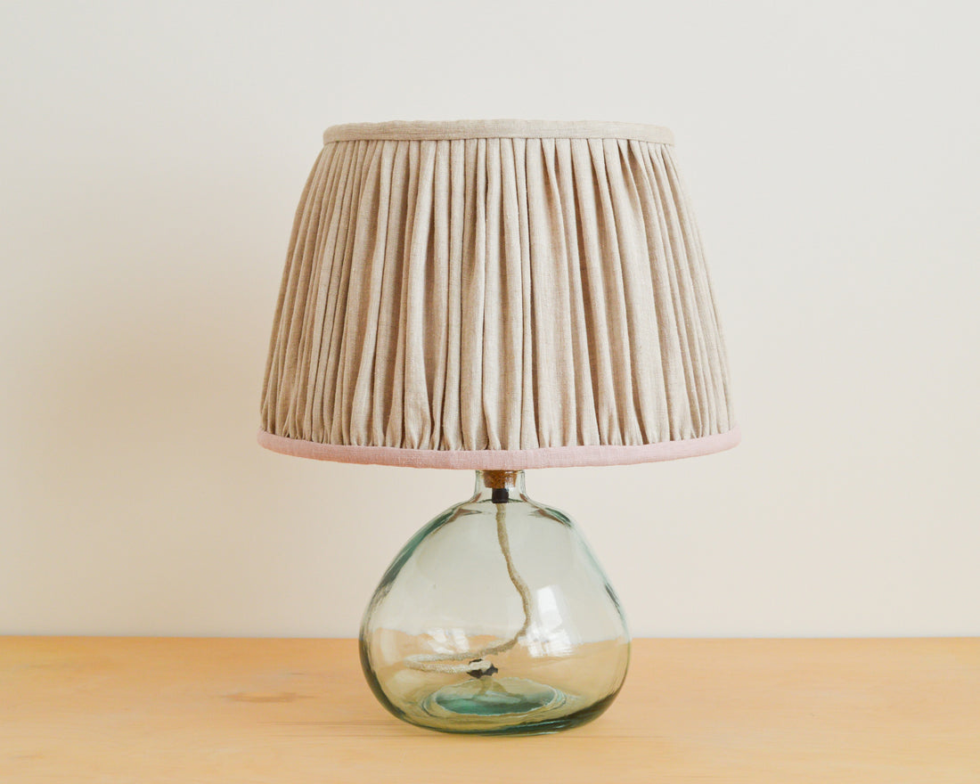 A natural linen gathered lampshade with a pink gathered texture, placed on a wooden surface with a glass lamp base.
