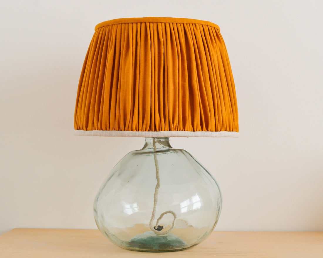 A mustard yellow gathered lampshade with a natural linen lining, placed on a glass lamp base.