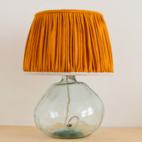 A mustard yellow gathered lampshade with a natural linen lining, placed on a glass lamp base.